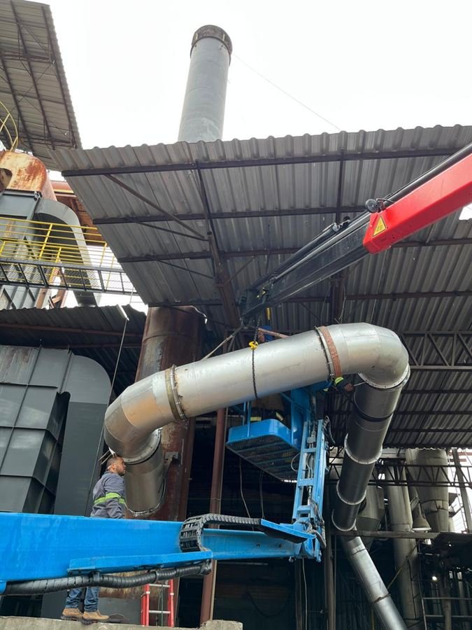 Duto metálico de ventilação sendo içado por guindaste e posicionado por montador em plataforma elevatória